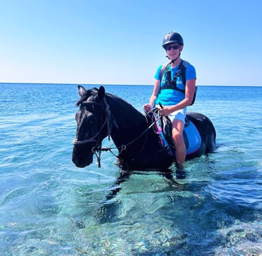 looneytravels riding a black horse through clear blue ocean water during a summer beach horse trek.