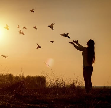 Mulher em pé em um por do sol, soltando pássaros no céu.