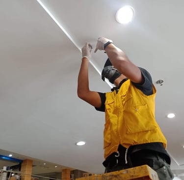 Electrician in a yellow safety vest installing recessed LED lighting in a white ceiling.