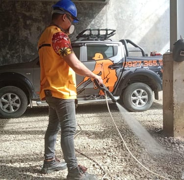 A worker using a high-pressure power washer to clean a gravel surface near a pickup truck.