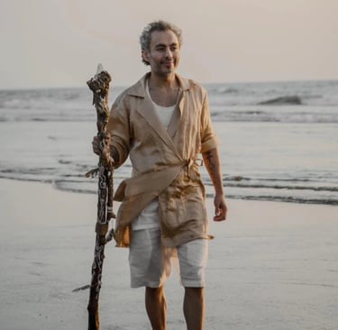 A man in linen clothing walks on a sandy beach holding a decorative wooden staff at sunset.