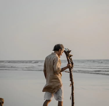 A man in linen clothing walking barefoot on a sandy beach at sunset holding a driftwood staff.