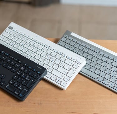Three wireless Bluetooth keyboards in black, white, and silver on a wooden desk.