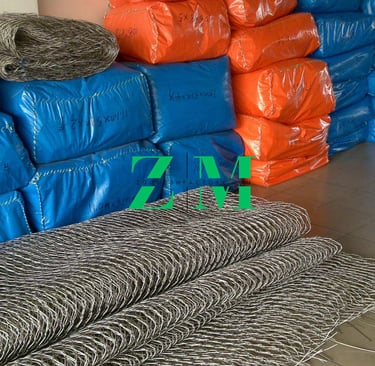 Rolls of flexible stainless steel wire rope mesh near blue and orange industrial packaging bags.