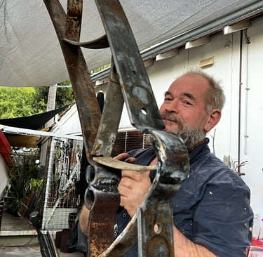A bearded metal artist working on a large, curved steel sculpture in an outdoor workshop.