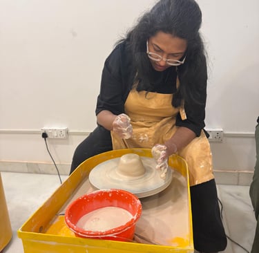 A woman wearing a yellow apron uses a pottery wheel to shape clay during a pottery workshop.