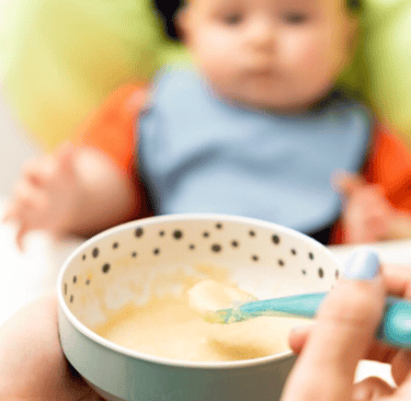 madre alimenta a un bebé con puré orgánico en una silla alta usando una cuchara azul.