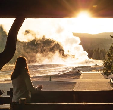 A woman watches the Old Faithful geyser erupt at sunset from a Yellowstone lodge porch.