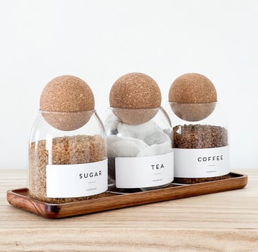 Minimalist glass canisters for sugar, tea, and coffee with round cork lids on a wooden tray.
