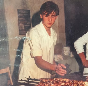 A vintage photo of a young chef in a white uniform grilling meat skewers on an open flame.