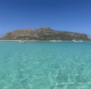 Crystal clear turquoise water at Balos Lagoon in Crete with boats near a rocky island.