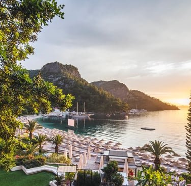 Sunset view over a luxury beach resort in a tropical cove with mountains, palm trees, and ocean boats.