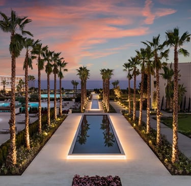Luxury tropical resort at sunset with lit reflecting pools, palm trees, and ocean views.