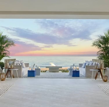 Luxury outdoor lounge with beach views, white sofas, and palm trees during a sunset at a coastal resort.