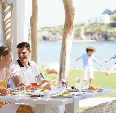A couple enjoys a luxury outdoor lunch at a beach resort while children play on the grass.