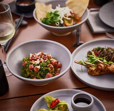 A gourmet spread of Asian fusion dishes including tuna tartare, chicken satay, and noodles served with white wine.