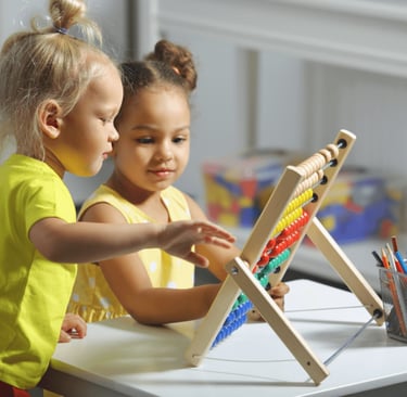 Two young girls learning math using a wooden abacus with colorful beads in a classroom.