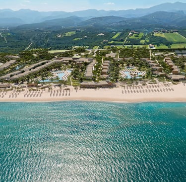 Aerial view of Ikos Kissamos resort with turquoise ocean water, sandy beach, and mountain backdrop.