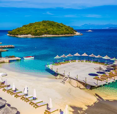 Tropical white sand beach in Ksamil, Albania, with straw umbrellas and a wooden deck overlooking turquoise waters.