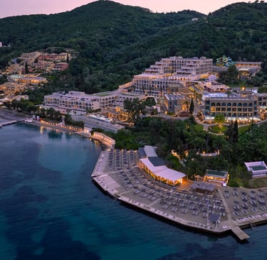 Aerial view of a luxury beachfront resort in Corfu at dusk with lighted pools and private beach.