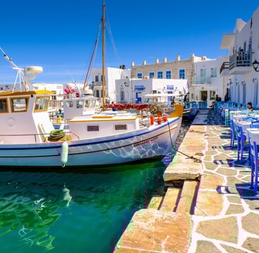 Traditional fishing boats docked by a waterfront Greek restaurant with blue chairs in Paros, Greece.