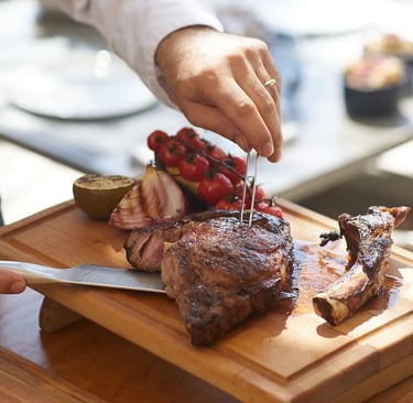 A chef carves a grilled ribeye tomahawk steak on a wooden board with roasted tomatoes and onions.