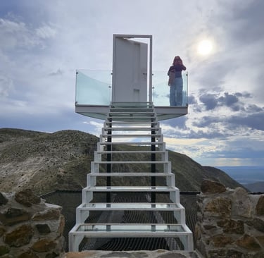 Una mujer se encuentra de pie en una escalera de cristal que conduce a una puerta blanca con vistas a un paisaje montañoso.