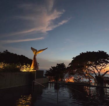 Piscina infinita de lujo al atardecer, con una escultura de cola de ballena y faroles luminosos.