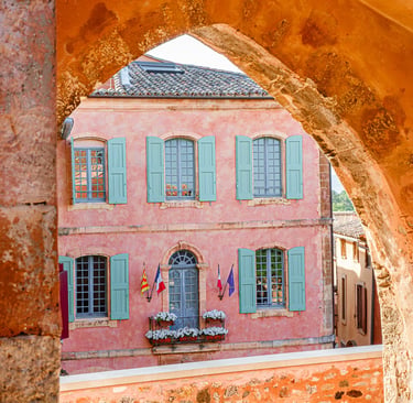 View of a pink Provencal building with teal shutters through a stone arch in Roussillon, France.