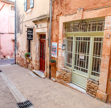 A charming narrow street in Roussillon, France, featuring terracotta buildings and an art gallery storefront.