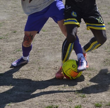 Due calciatori lottano per il possesso di un pallone giallo-arancione su un campo in terra, con le g