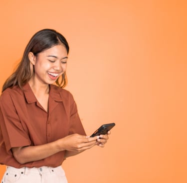 Smiling young woman holding a smartphone and laughing while texting on an orange background.