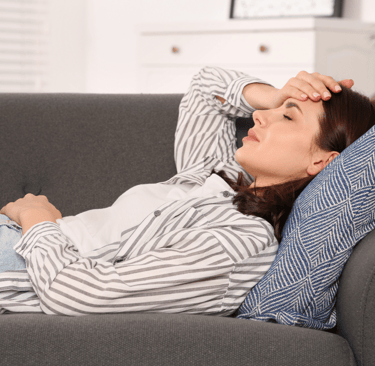 woman laying on a couch with one hand on head and one on stiomach, seems to be in pain