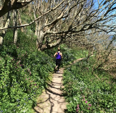A woman trail running on a sunny path through a lush green forest with blooming wildflowers.