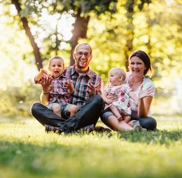 homem e mulher sentados em um gramado, cada um segura uma criança, expressão de felicidade no rosto