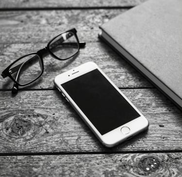 Black and white flat lay of a smartphone, eyeglasses, and notebook on a rustic wooden desk.