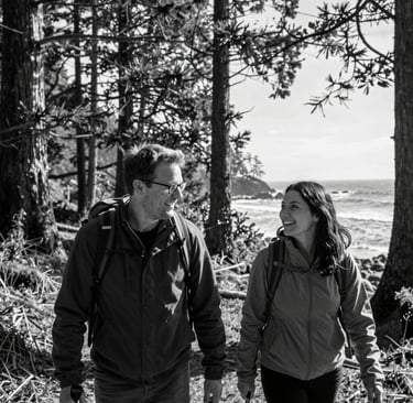 Smiling couple hiking through a coastal forest with ocean views and tall redwood trees.