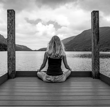 A woman practices yoga meditation on a wooden pier overlooking a scenic mountain lake.