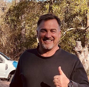 A smiling man with a goatee giving a thumbs up outdoors with trees and a truck in the background.