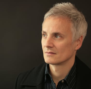 Professional portrait of a man with short grey hair and green eyes looking aside against a dark background.
