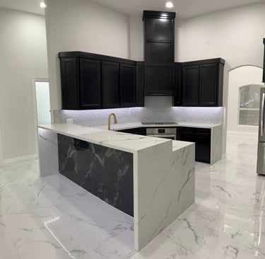 Modern kitchen featuring black cabinets, white marble waterfall island, and gold faucet fixtures.