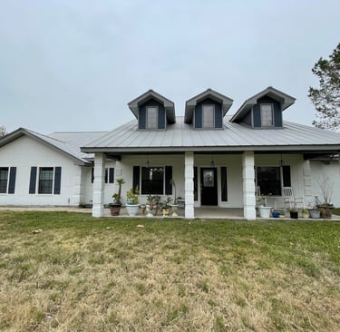 White brick ranch style home with a grey metal roof and three dormer windows on a grassy lawn.