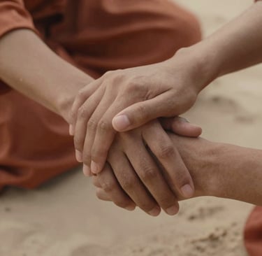 A close-up, authentic shot of two hands gently clasped together, capturing a moment of genuine human connection. The scene is bathed in a warm, inviting glow with terracotta-colored fabrics in the background and soft sand skin tones.