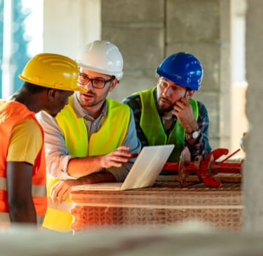 Three construction engineers wearing hard hats and safety vests discuss project blueprints on a laptop at a building site.