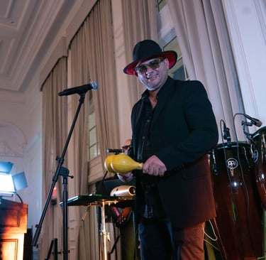 A male musician in a black suit and hat plays yellow maracas with conga drums at a live event.