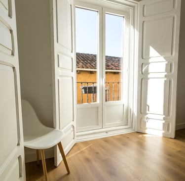Minimalist room with light oak wood floors, white shutters, and a modern chair by a sunlit window.