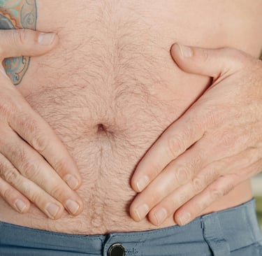 Close‑up of an adult male abdomen with hands resting on the stomach, showing natural body hair and a side tattoo, outdoors