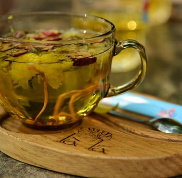 “Glass cup of herbal tea with visible flower blossoms on a wooden saucer, placed beside a spoon and a blue packet 