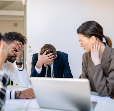 Four people sit around a table in an office meeting, with three of them resting their heads in their hands