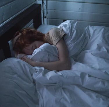 A redhead woman sleeping deeply in a dark bedroom with white bedding and a wooden bed frame.